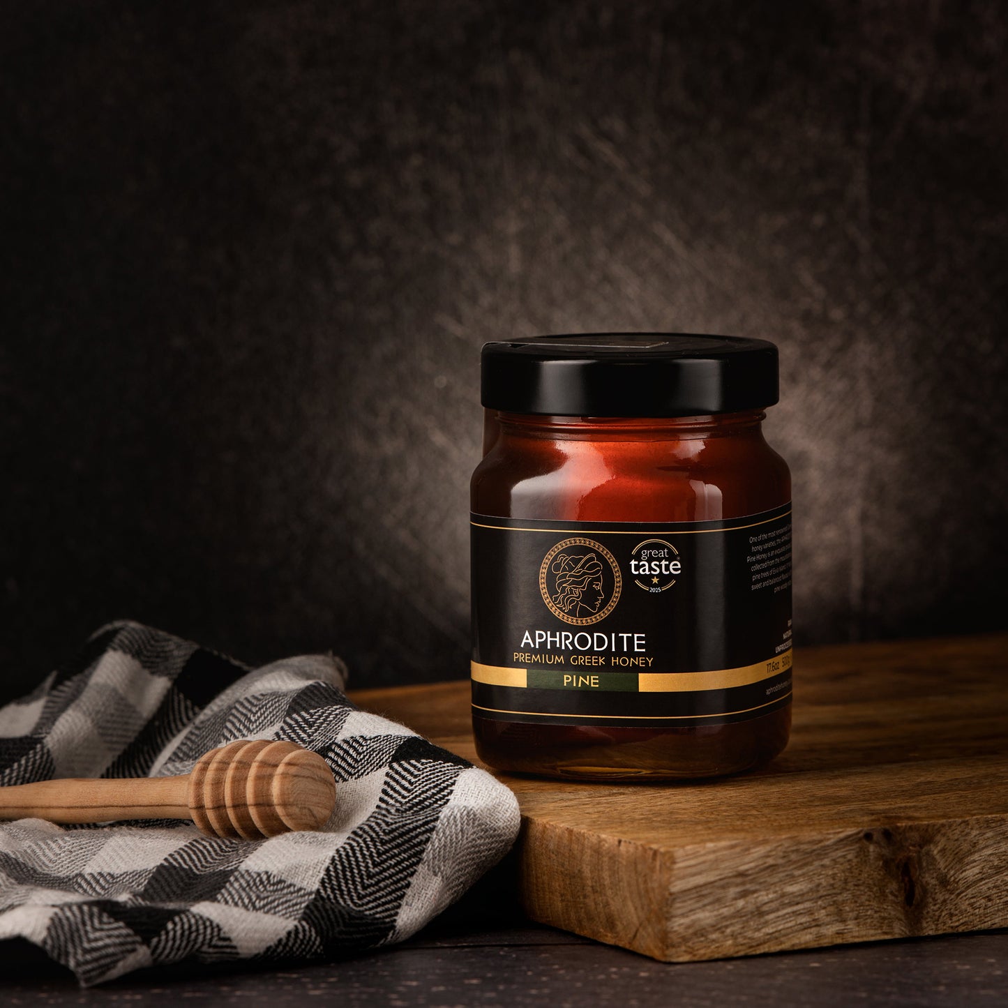 Aphrodite Raw Pine Honey 480g jar on a wooden board with a checkered cloth and honey dipper, dark textured background