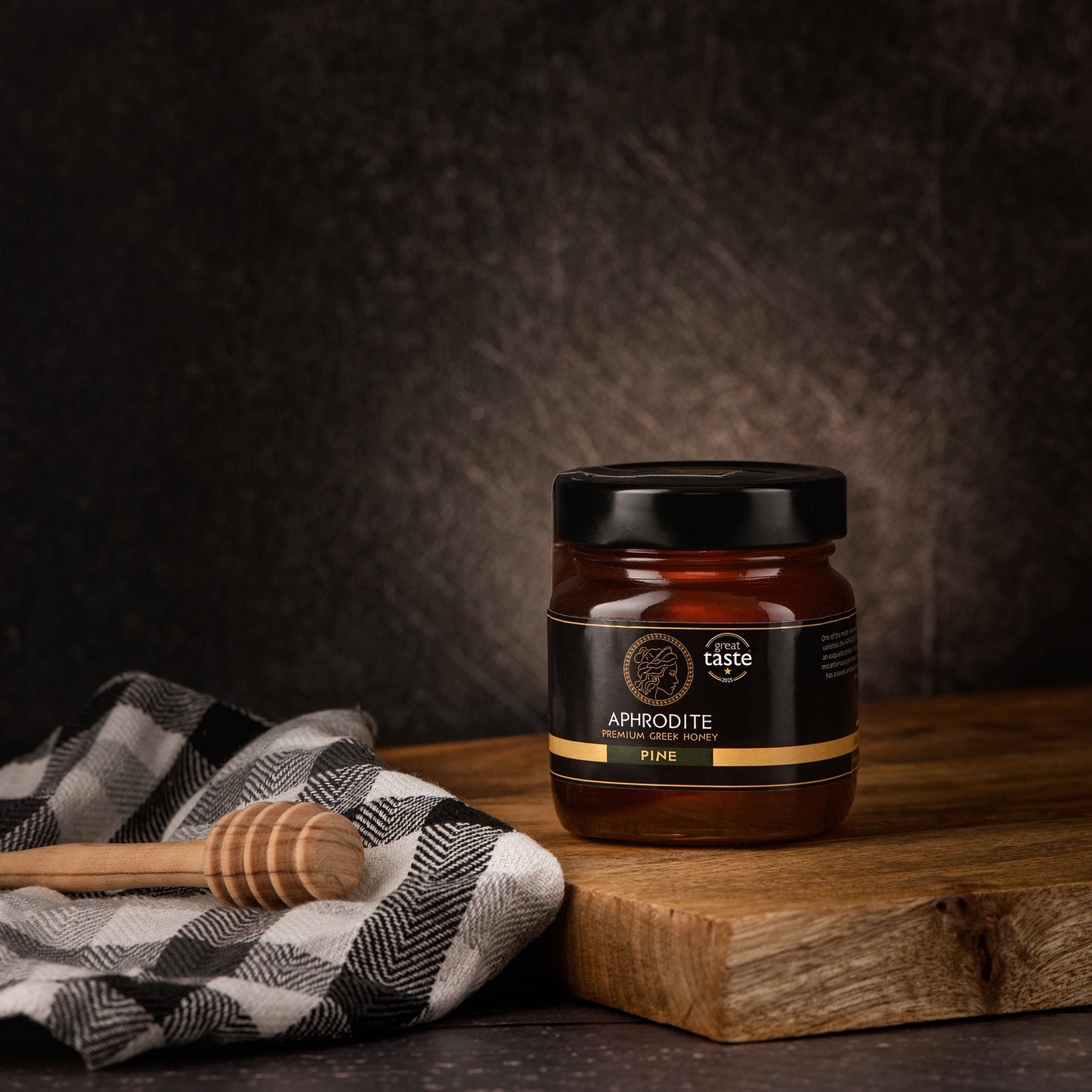 Aphrodite Raw Pine Honey 250g jar on a wooden board with a checkered cloth and honey dipper, dark textured background