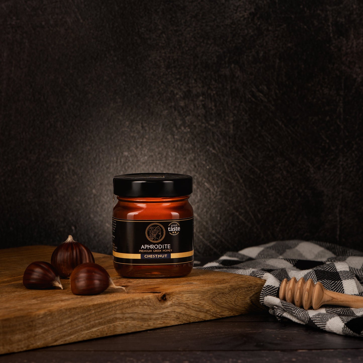 Greek Raw Chestnut Honey in a 250g glass jar in dark / black background. Jar presented from the front . Chestnuts on the side. 100% Pure Honey. Aphrodite Honey. Aphrodite Premium Greek Honey. Great Taste 2 stars.