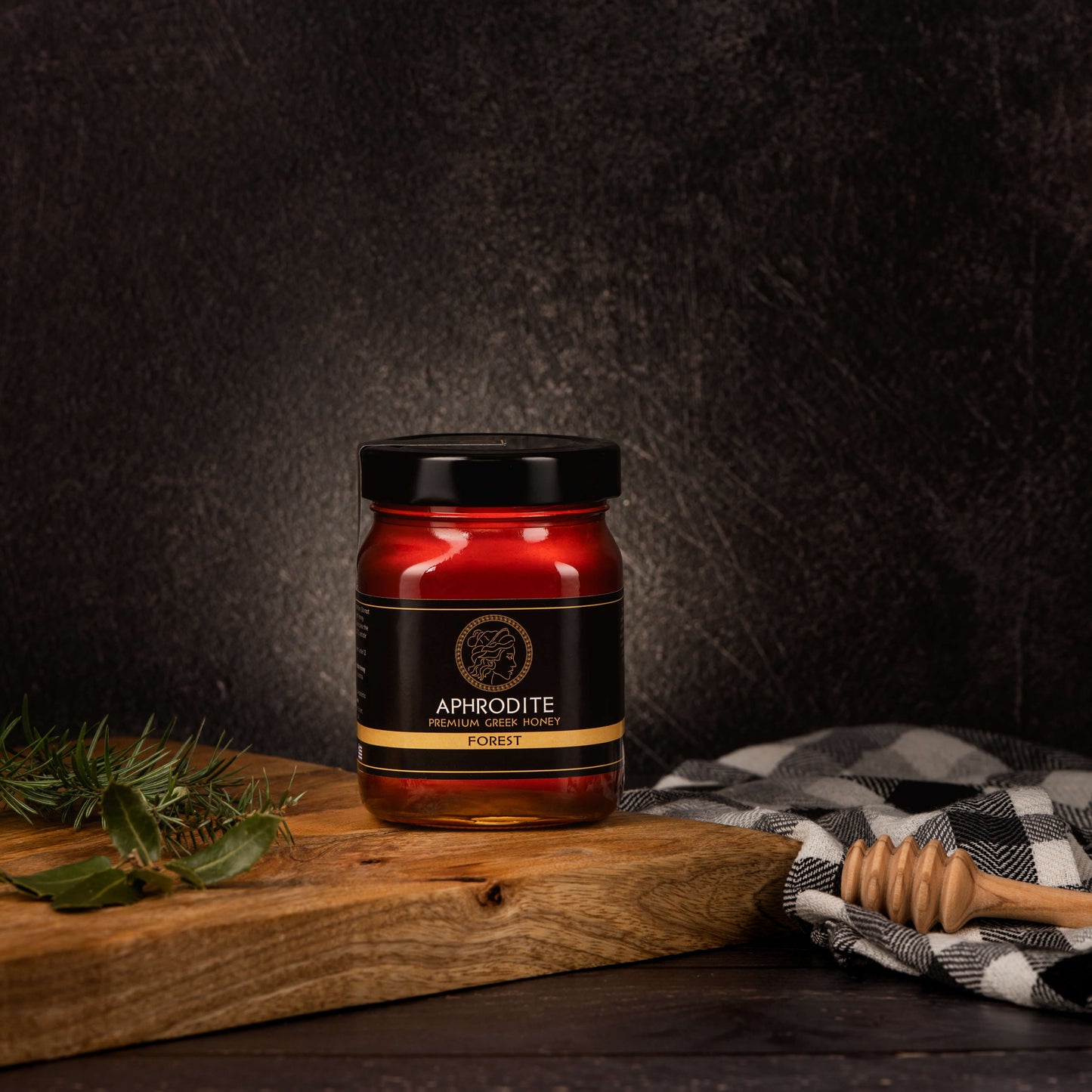 Greek Raw Forest Honey in a 500g glass jar in dark / black background. Jar presented from the front . Tree leaves on the side. 100% Pure Honey. Aphrodite Honey. Aphrodite Premium Greek Honey.