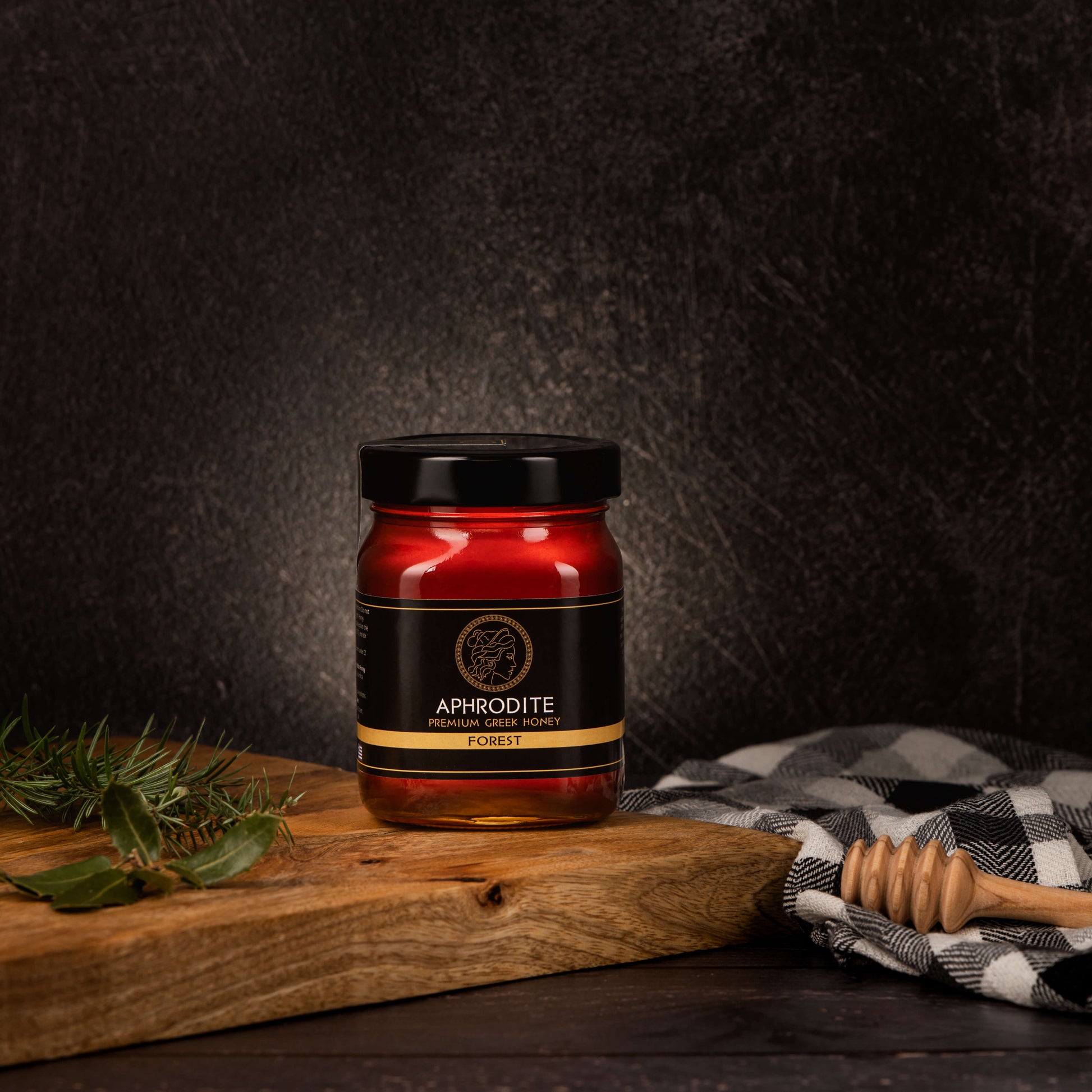 Greek Raw Forest Honey in a 500g glass jar in dark / black background. Jar presented from the front . Tree leaves on the side. 100% Pure Honey. Aphrodite Honey. Aphrodite Premium Greek Honey.