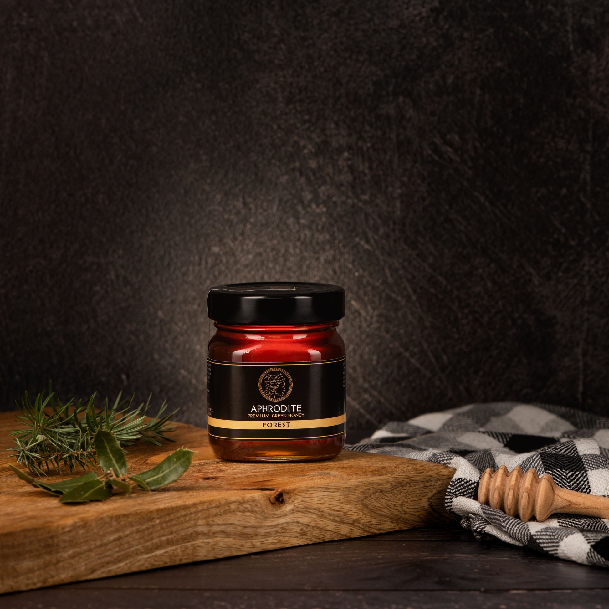 Greek Raw Forest Honey in a 250g glass jar in dark / black background. Jar presented from the front . Tree leaves on the side. 100% Pure Honey. Aphrodite Honey. Aphrodite Premium Greek Honey.