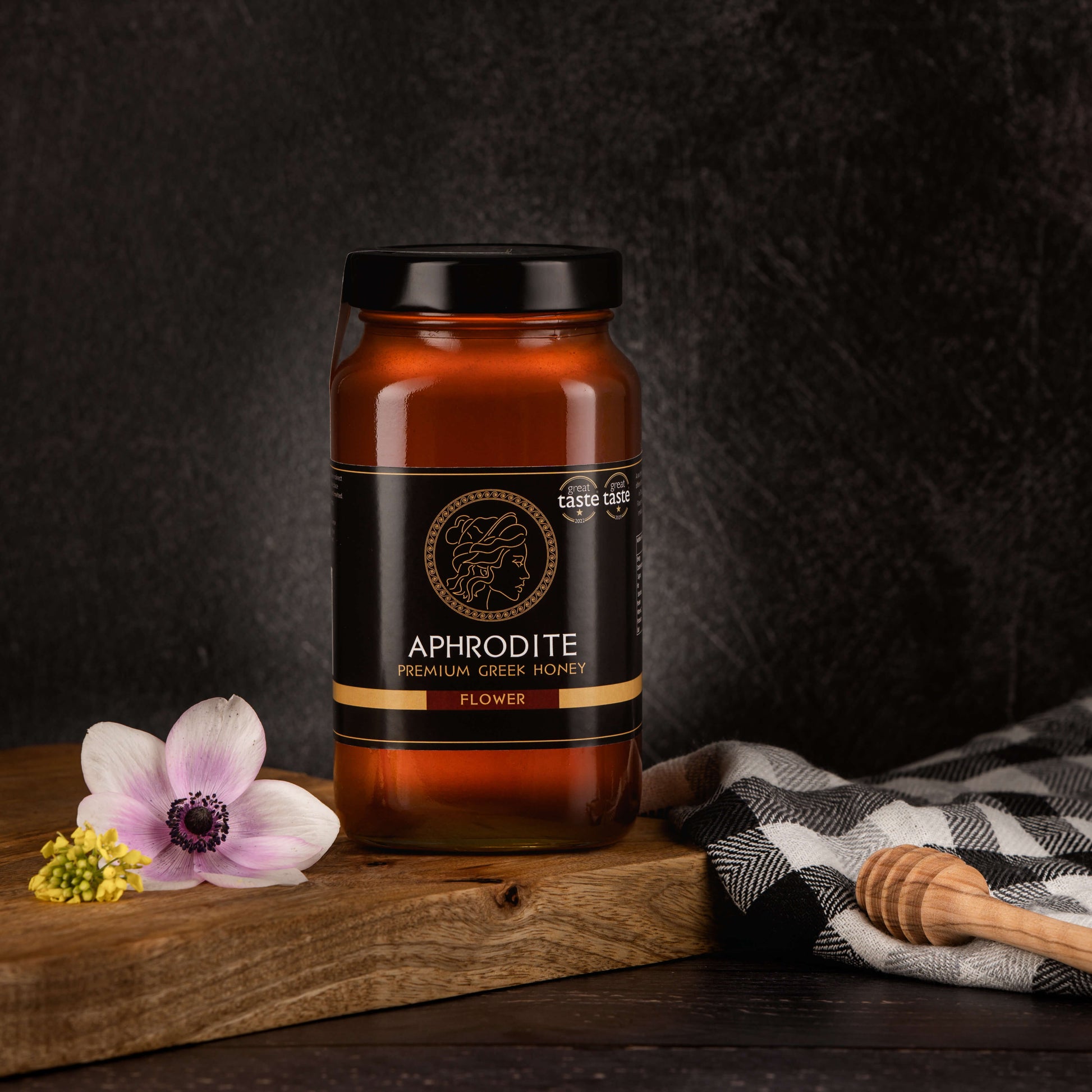 Greek Raw Flower / Blossom Honey in a 950g glass jar in dark / black background. Jar presented from the front . Flowers on the side. 100% Pure Honey. Aphrodite Honey. Aphrodite Premium Greek Honey. Great Taste 1 star.