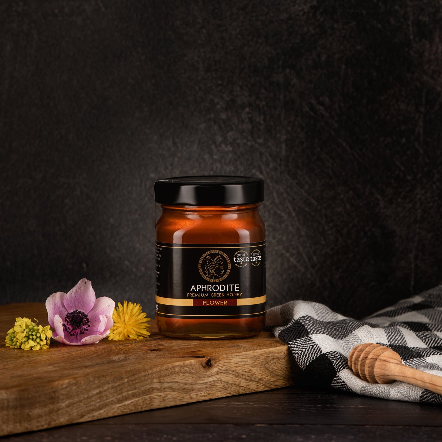 Greek Raw Flower / Blossom Honey in a 500g glass jar in dark / black background. Jar presented from the front . Flowers on the side. 100% Pure Honey. Aphrodite Honey. Aphrodite Premium Greek Honey. Great Taste 1 star.