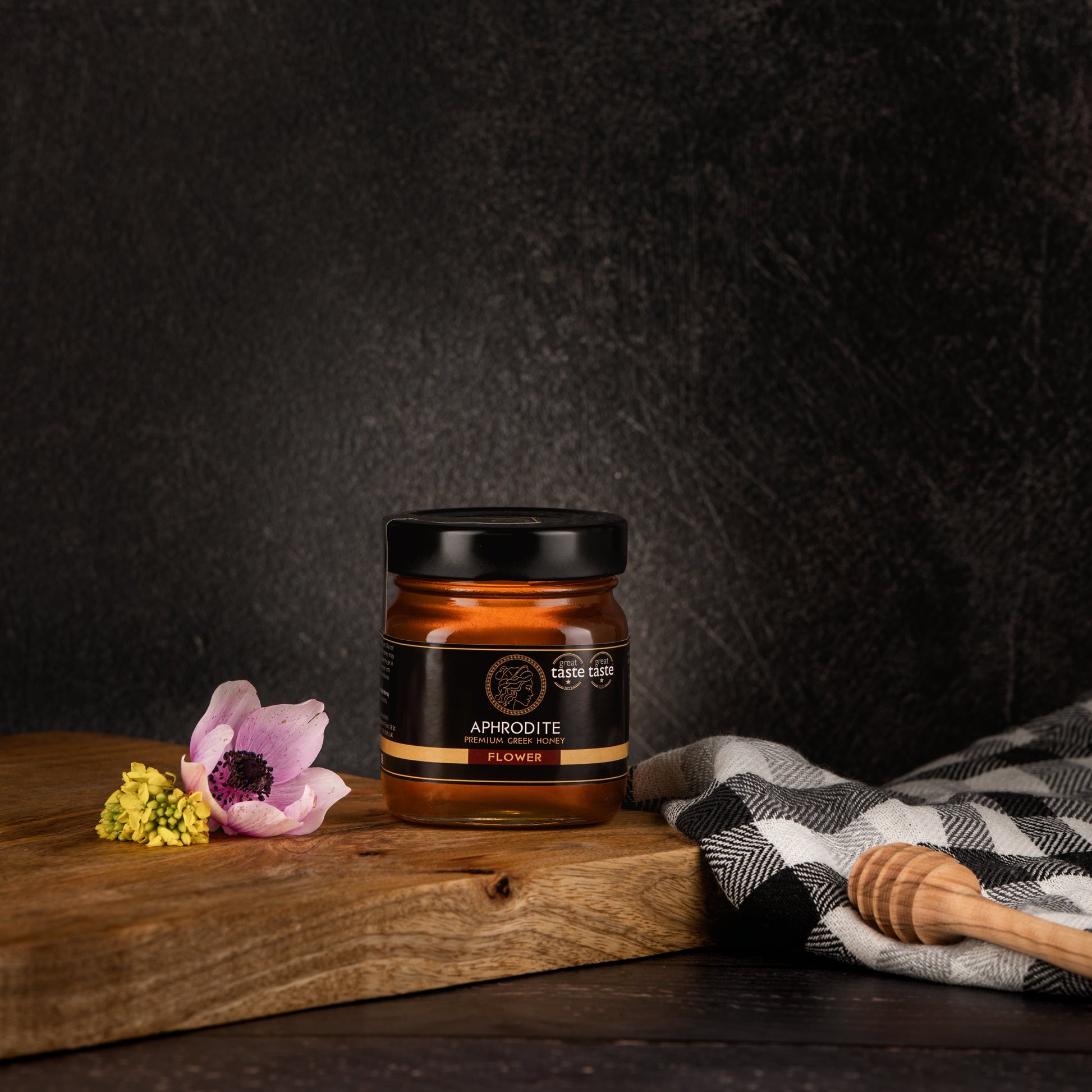 Greek Raw Flower / Blossom Honey in a 250g glass jar in dark / black background. Jar presented from the front . Flowers on the side. 100% Pure Honey. Aphrodite Honey. Aphrodite Premium Greek Honey. Great Taste 1 star.