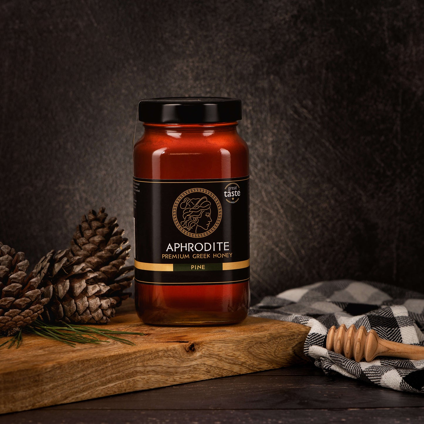 Aphrodite Raw Pine Honey 950g jar on a rustic wooden board with pinecones, a checkered cloth and honey dipper, dark textured background