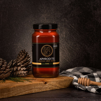 Aphrodite Raw Pine Honey 950g jar on a rustic wooden board with pinecones, a checkered cloth and honey dipper, dark textured background