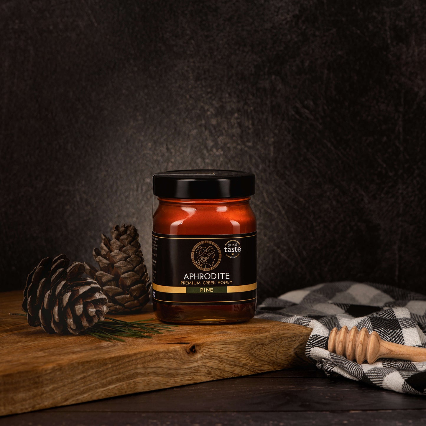 Aphrodite Raw Pine Honey 480g jar on a rustic wooden board with pinecones, a checkered cloth and honey dipper, dark textured background