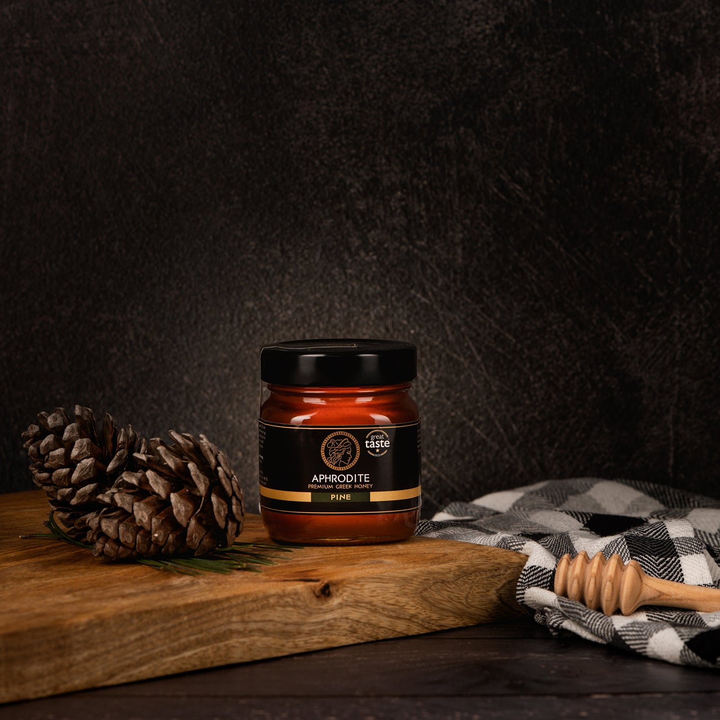 Aphrodite Raw Pine Honey 250g jar on a rustic wooden board with pinecones and a honey dipper, dark textured background