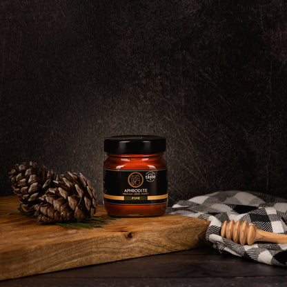 Aphrodite Raw Pine Honey 250g jar on a rustic wooden board with pinecones and a honey dipper, dark textured background