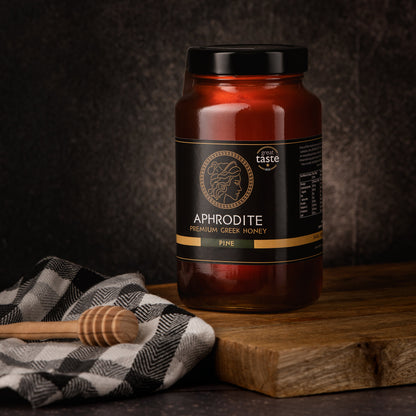 Aphrodite Raw Pine Honey 950g jar on a wooden board with a checkered cloth and honey dipper, dark textured background