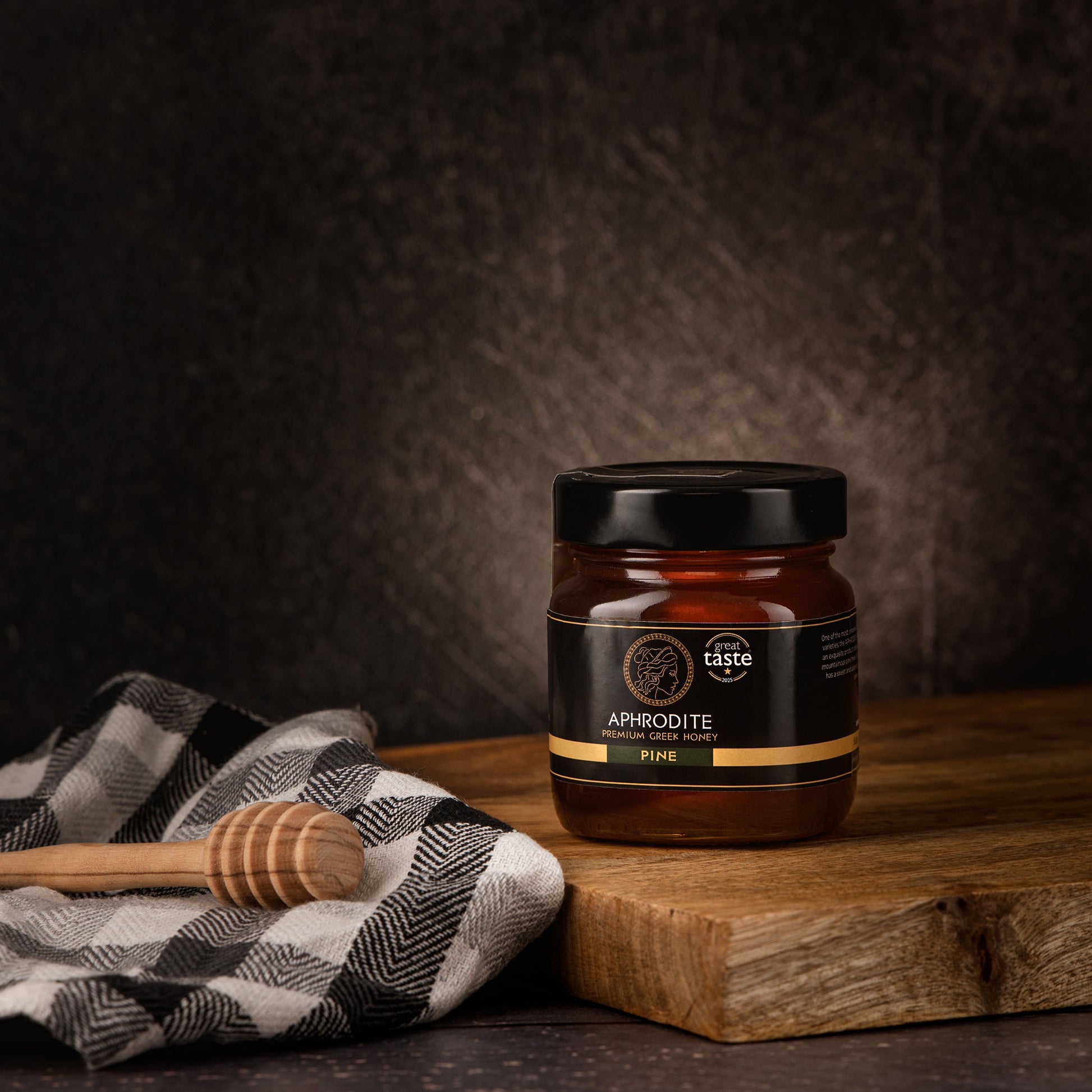 Aphrodite Raw Pine Honey 250g jar on a wooden board with a checkered cloth and honey dipper, dark textured background