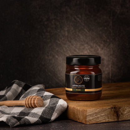 Aphrodite Raw Pine Honey 250g jar on a wooden board with a checkered cloth and honey dipper, dark textured background