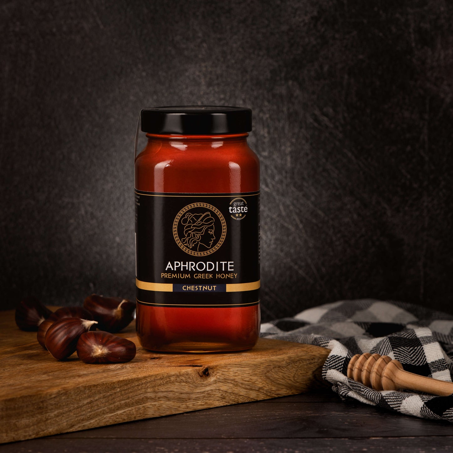 Greek Raw Chestnut Honey in a 950g glass jar in dark / black background. Jar presented from the front . Chestnuts on the side. 100% Pure Honey. Aphrodite Honey. Aphrodite Premium Greek Honey. Great Taste 2 stars.