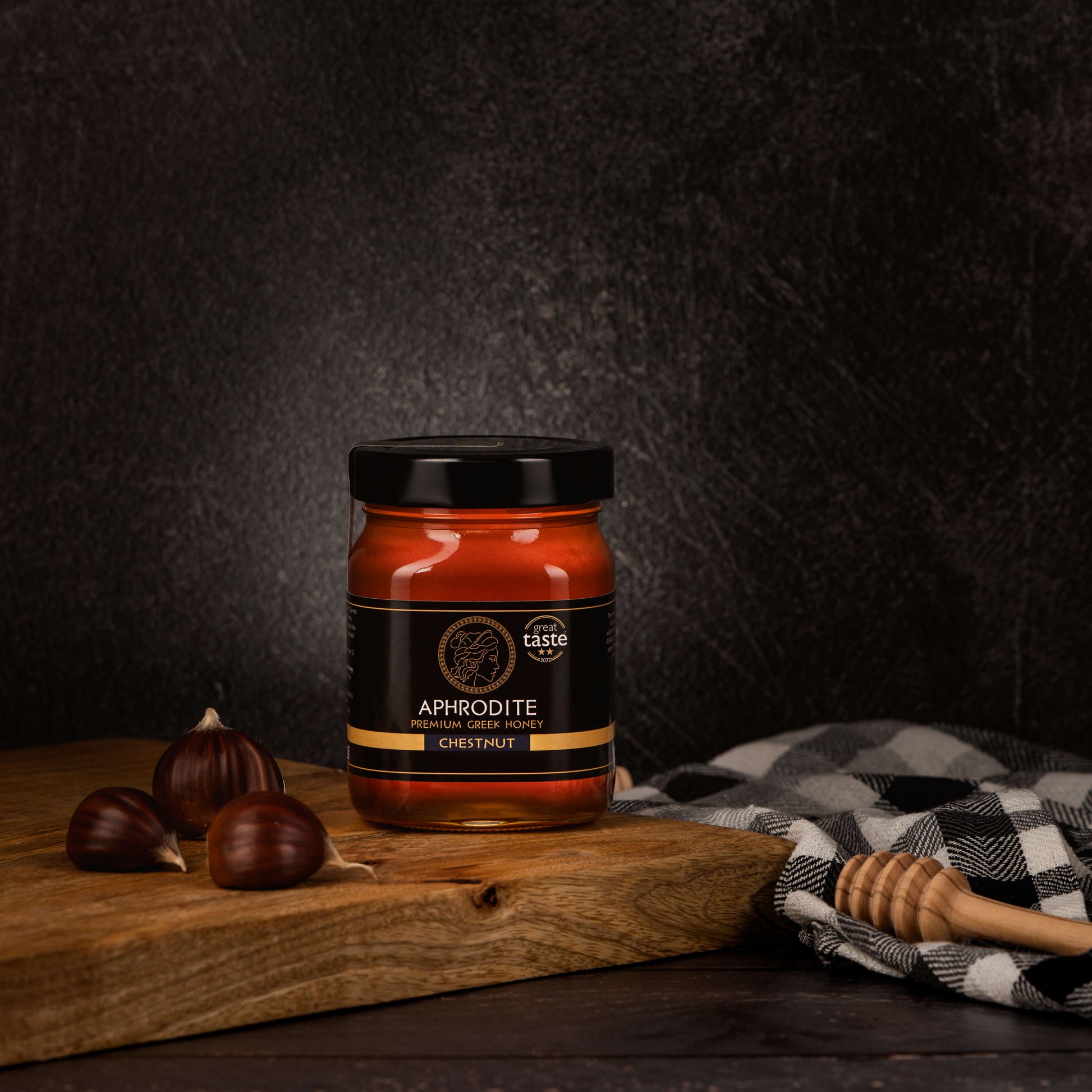 Greek Raw Chestnut Honey in a 500g glass jar in dark / black background. Jar presented from the front . Chestnuts on the side. 100% Pure Honey. Aphrodite Honey. Aphrodite Premium Greek Honey. Great Taste 2 stars.