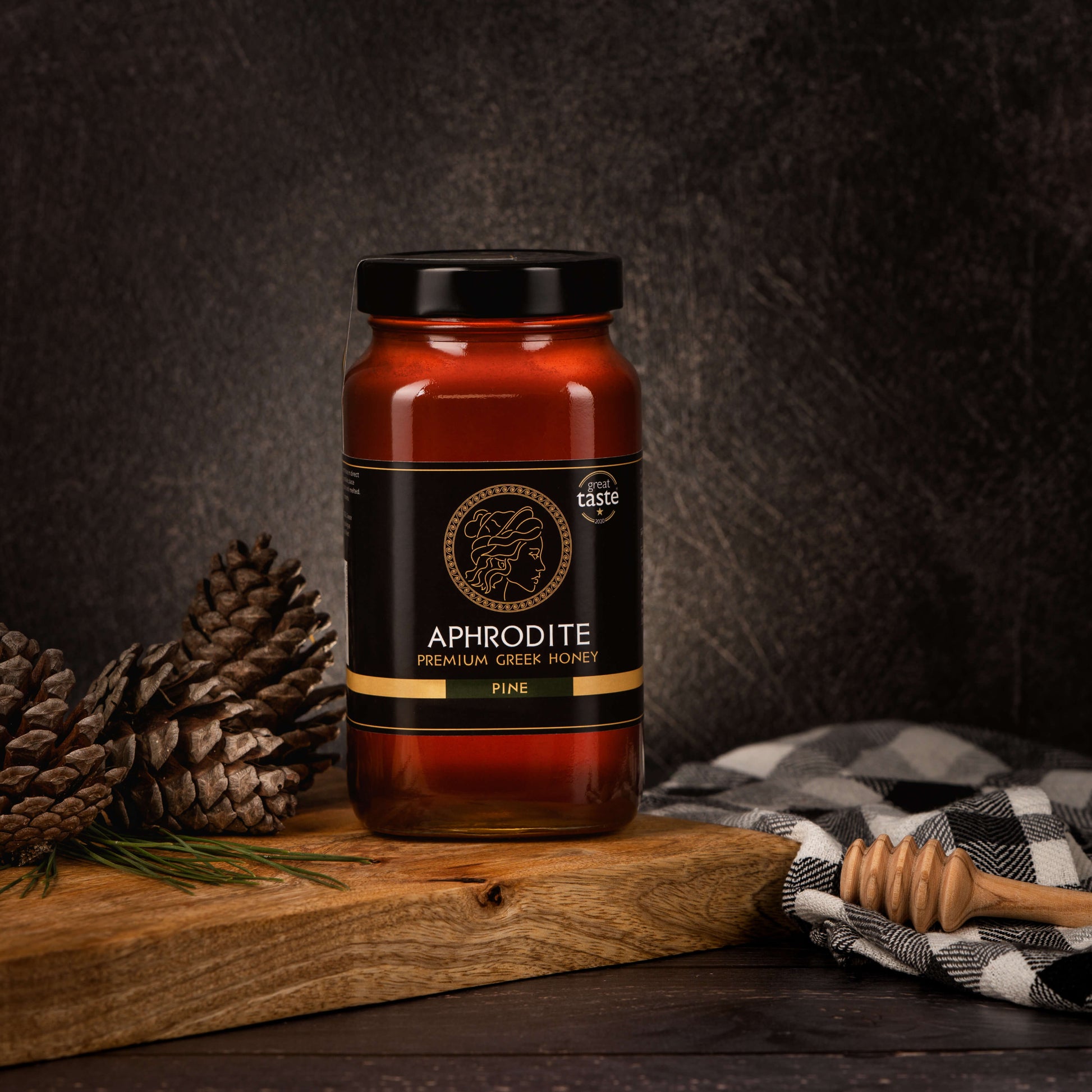 Greek Raw Pine Honey in a 950g glass jar in dark / black background. Jar presented from the front . Pine cones on the side. 100% Pure Honey. Aphrodite Honey. Aphrodite Premium Greek Honey. Great Taste 1 star.