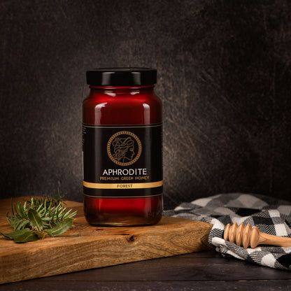 Greek Raw Forest Honey in a 950g glass jar in dark / black background. Jar presented from the front . Tree leaves on the side. 100% Pure Honey. Aphrodite Honey. Aphrodite Premium Greek Honey.